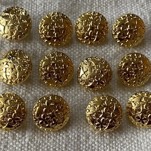 Puede incluir: Doce botones dorados con un diseño floral. Los botones son redondos y tienen un patrón en relieve texturizado. Los botones están dispuestos en tres filas de cuatro sobre una superficie blanca texturizada.