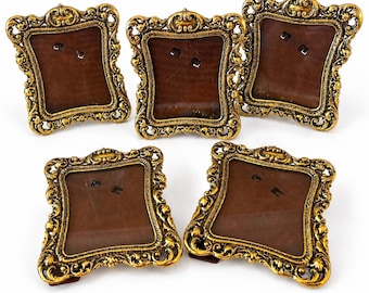 Può includere: Un set di cinque cornici rettangolari con bordi in metallo dorato in rilievo, caratterizzate da intricati motivi a volute e dettagli a forma di foglia. Ogni cornice presenta un retro marrone scuro e un pannello frontale in vetro.