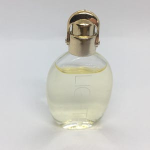 Könnte beinhalten: Eine kleine, klare Glasparfümflasche mit goldfarbenem Verschluss und gebogenem Griff. Die Flasche enthält eine hellgelbe Flüssigkeit und trägt den Aufdruck "LIG" auf der Vorderseite. Die Flasche hat eine runde Form.