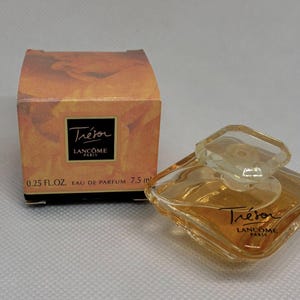 Könnte beinhalten: Ein kleiner Flakon des Parfums Lancôme Trésor. Die klare, geometrisch geformte Flasche hat einen passenden Deckel und enthält eine goldfarbene Flüssigkeit. Die Schachtel ist orange und trägt das Trésor-Logo.