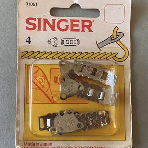 Könnte beinhalten: Eine Packung mit vier Metallverschlüssen der Marke Singer für Röcke und Kleider. Die Verpackung ist gelb und weiß mit dem Singer-Logo und Produktinformationen. Die Verschlüsse sind silberfarben und haben ein dekoratives Design.