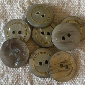 Peut inclure: Un ensemble de huit gros boutons ronds de différentes nuances de gris. Chaque bouton comporte deux trous et un anneau doré sur le bord. Les boutons sont disposés sur une surface texturée blanc cassé.