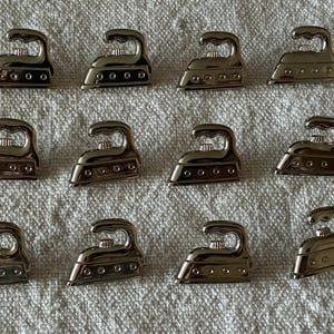 Pode incluir: Uma coleção de pequenos pingentes prateados em forma de ferro de passar. Os pingentes estão dispostos sobre uma superfície texturizada em branco sujo. Cada pingente tem uma alça e uma base detalhada com pequenas reentrâncias circulares.