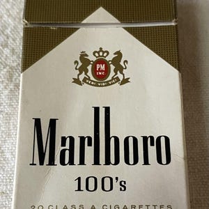 Pode incluir: Um maço de cigarros Marlboro 100's. O maço é branco com uma parte superior dourada e castanha. O logotipo da Marlboro é preto, com o texto "100's" abaixo. A parte superior do maço diz "FILTER CIGARETTES".