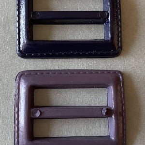 Pode incluir: Duas fivelas quadradas, uma azul marinho escuro e outra roxa suave. Ambas têm formato retangular com uma barra central e um pequeno pino. As bordas são arredondadas e parecem costuradas. Adequadas para projetos de artesanato.