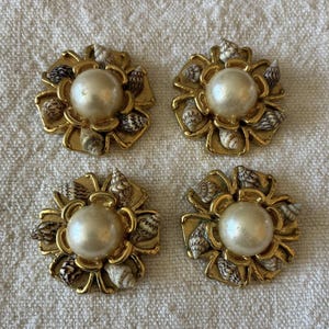 May include: Four gold-coloured flower-shaped earrings. Each earring has a large, round, off-white pearl in the centre, surrounded by small seashells and gold-coloured petals. The earrings are arranged on a textured, off-white surface.