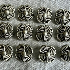May include: A collection of twelve silver-colored, flower-shaped buttons. Each button has a central, raised circle surrounded by detailed, radiating petals. The buttons are arranged on a textured, light gray fabric.