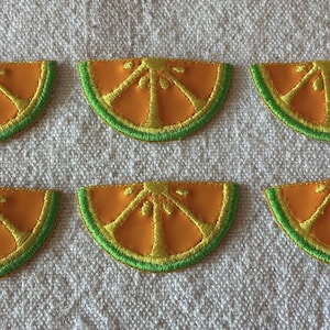 Pode incluir: Seis patches bordados de fatias de laranja. Cada patch apresenta um centro laranja brilhante com detalhes amarelos e uma casca verde. Os patches estão dispostos sobre um tecido texturizado branco sujo.