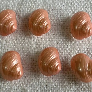 Pode incluir: Seis contas peroladas em forma de concha, cor pêssego, dispostas sobre uma superfície branca texturizada. Cada conta tem um desenho em espiral que capta a luz. Estas contas decorativas podem ser usadas para fazer joias ou outros projetos de artesanato.