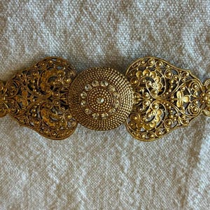 May include: A gold-toned metal hair barrette featuring an intricate filigree design with scrolling patterns on both sides and a central circular medallion. The medallion is textured with small raised dots and embedded with clear, sparkling rhinestones.