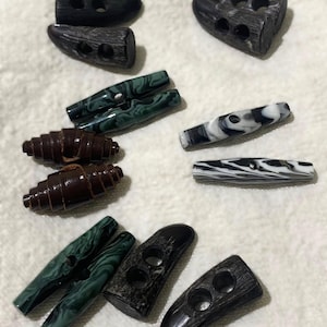 Pode incluir: Uma coleção de botões de alternância em vários estilos e cores. Os botões incluem designs marmorizados em marrom escuro, preto, verde e branco. Alguns têm dois orifícios, outros três. Os botões são feitos de madeira e outros materiais.