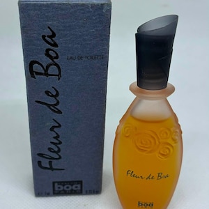 Può includere: Una boccetta di Eau de Toilette Fleur de Boa con corpo in vetro satinato arancione con dettagli a rosa in rilievo e tappo grigio scuro, accompagnata da una scatola alta, rettangolare e testurizzata blu scuro.