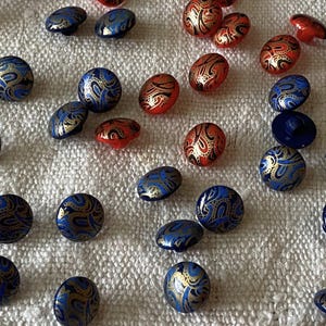 Könnte beinhalten: Eine Auswahl an dekorativen Knöpfen in verschiedenen Formen. Die Knöpfe weisen ein blaues, goldenes und rotes Paisley-Muster auf. Einige sind rund, andere oval oder gewölbt. Die Knöpfe sind auf einer weißen, strukturierten Oberfläche verstreut.