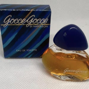 May include: A miniature perfume bottle with a dark blue cap and a clear, rounded body containing amber liquid. Beside it is a box with the words "Gocce Gocce di Napoleon" and "Eau de Toilette" printed on it.