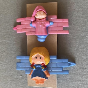 Op de afbeelding: Twee vintage plastic beeldjes. De bovenste figuur is een roze gekleed figuur met een bijpassende hoed en blauwe accenten, bevestigd aan een roze bakstenen muur. De onderste figuur is een blondharige figuur in een blauwe jurk, bevestigd aan een blauwe bakstenen muur.
