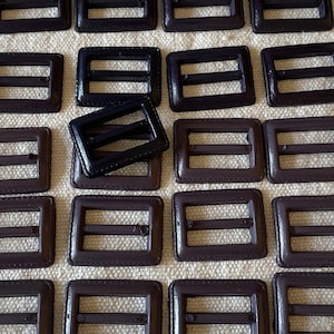 May include: A collection of dark brown rectangular buckles with a central bar. The buckles are arranged on a textured, off-white surface. The buckles are made of a smooth, possibly leather-like material, and are evenly spaced.