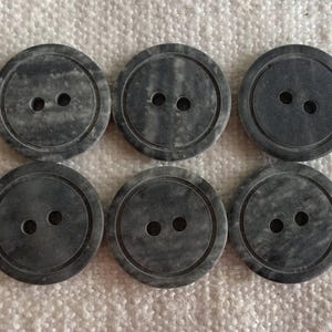 Pode incluir: Seis botões redondos cinza escuro com dois furos cada. Os botões têm uma aparência marmorizada e um anel externo em relevo. Estão dispostos em duas fileiras de três em uma superfície branca texturizada.