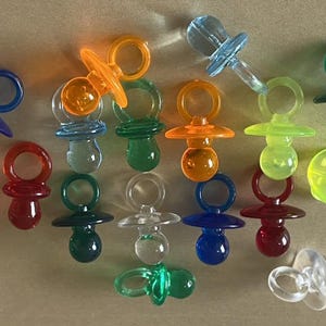Pode incluir: Uma coleção de chupetas coloridas para bebês em várias tonalidades, incluindo azul, laranja, verde, vermelho e transparente. As chupetas são feitas de plástico transparente e estão dispostas sobre um fundo neutro. A imagem mostra uma variedade de cores e designs.