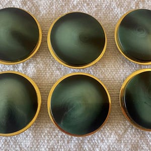Puede incluir: Seis posavasos redondos de color verde oscuro con borde dorado. Los posavasos tienen un diseño en espiral en el centro. Los posavasos están dispuestos en dos filas de tres sobre una superficie blanca texturizada.