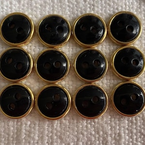 Peut inclure: Douze boutons noirs avec des bordures dorées. Chaque bouton comporte deux trous pour la couture. Les boutons sont disposés en trois rangées de quatre boutons chacun. Les boutons sont sur une surface blanche et texturée.