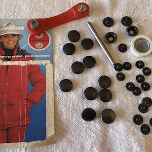 May include: A collection of black press fasteners, a red tool, and a silver tool are scattered on a white surface. A package of Prym brand fasteners is also present, featuring a woman in a red jacket. The package reads "10 Druckknöpfe boutons-pression press fasteners."