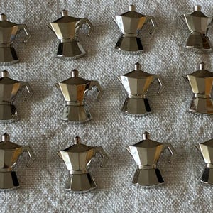 Puede incluir: Una colección de doce pequeñas cafeteras de espresso plateadas dispuestas en un patrón de cuadrícula. Cada cafetera en miniatura presenta un diseño clásico con un asa y una tapa, lo que refleja una cafetera italiana tradicional.