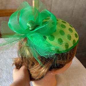 Può includere: Un fascinator verde brillante caratterizzato da una base rotonda con un motivo a pois verde scuro. Il cappello è decorato con un grande fiocco a strati in rete verde trasparente e un elemento centrale circolare, per un aspetto voluminoso.