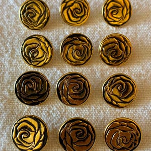May include: A set of twelve circular buttons arranged in four rows of three. Each button features a raised, three-dimensional rose design in a metallic gold finish with dark, recessed detailing that highlights the petal contours and shapes.