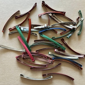 Pode incluir: Uma coleção de grampos de cabelo de metal em várias cores, incluindo prata, bordô, verde, rosa e azul. Os grampos são curvos e têm um design simples e funcional. A imagem é tirada de cima.