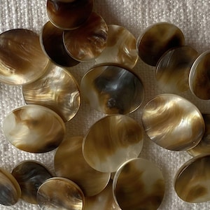 Può includere: Una collezione di bottoni rotondi e iridescenti nei toni del marrone, beige e crema. I bottoni hanno un motivo a vortice e marmorizzato e una finitura lucida. Sono sparsi su una superficie strutturata bianco sporco.