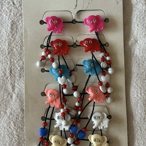 Op de afbeelding: Een kaart met kleurrijke haaraccessoires met olifantvormige bedels. De kaart bevat roze, rode, blauwe, witte en gele olifanten. Elke bedel is bevestigd aan een zwart elastiek met decoratieve kralen. De kaart heeft het woord "pasador" in het rood.