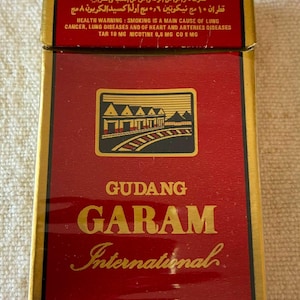 May include: A rectangular cigarette pack with a red and gold color scheme. The front features a graphic of a building with a roof, the text GUDANG GARAM International, and a health warning stating smoking is a main cause of lung cancer.
