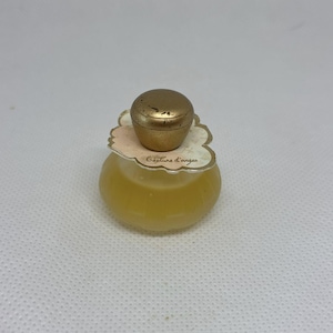 Pode incluir: Um pequeno frasco de perfume vintage com uma tampa dourada e um líquido amarelo pálido no interior. O frasco tem uma forma arredondada e um rótulo decorativo com o texto "Gesture d'anges". A tampa está ligeiramente desgastada.
