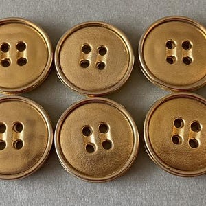 May include: Six gold-colored buttons with a round shape and four holes each. The buttons have a slightly raised rim and a matte finish. These buttons are suitable for sewing projects.