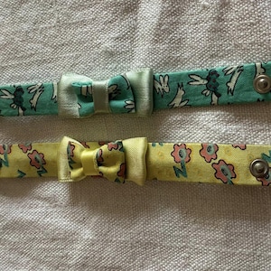 Pode incluir: Duas pulseiras de tecido com fechos de pressão e laços. Uma é verde-azulado com um padrão de coelhos brancos, a outra é amarela com um padrão floral rosa. Ambas têm um pequeno laço no centro.