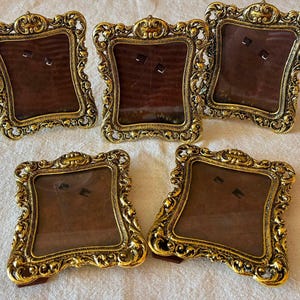 Può includere: Un set di cinque cornici rettangolari con bordi in metallo dorato ornati e in rilievo, caratterizzati da intricati motivi a volute e floreali. Ogni cornice presenta un retro marrone scuro e un frontale in vetro trasparente da tavolo.