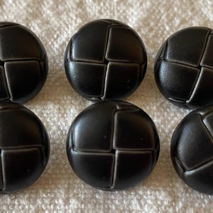 Pode incluir: Seis botões redondos pretos com um design segmentado e texturizado. Cada botão tem um padrão em forma de cruz criado pelos segmentos que se cruzam. Os botões estão dispostos sobre uma superfície branca e texturizada.
