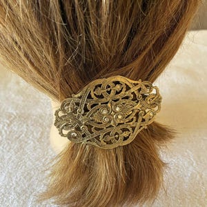 Könnte beinhalten: Verziertes goldfarbenes Haar-Accessoire mit einem aufwendigen Filigranmuster. Der Clip, verziert mit kleinen Kristallen, hält einen Teil hellbraunen Haares zurück. Dieses Haar-Accessoire bietet eine Vintage-Ästhetik.