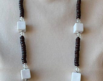 May include: A long necklace featuring dark brown disc-shaped beads alternating with white cube-shaped beads and small clear spherical beads. The beads are arranged in a repeating pattern along the length of the necklace, creating a geometric design.