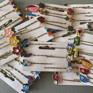 May include: A collection of decorative bobby pins with various colorful designs. The pins feature flower, animal, and cartoon character embellishments. Each pin is attached to a white card, ready for display or sale.