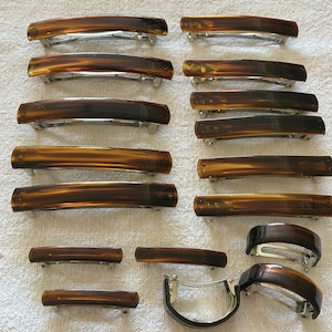 May include: A collection of tortoiseshell hair barrettes in various sizes and shapes. The barrettes have a glossy finish and silver-tone metal clasps. The colors range from light amber to dark brown.