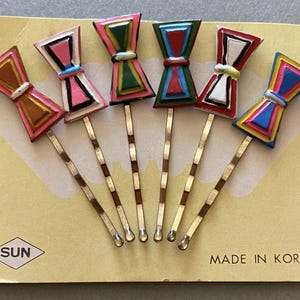 Può includere: Sei forcine per capelli vintage a forma di fiocco in vari colori, tra cui rosa, giallo, verde e blu, sono disposte su una carta gialla. Le forcine hanno steli in metallo color oro e sono etichettate "MADE IN KOREA".