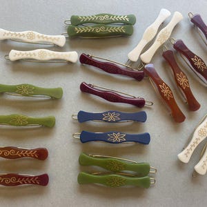 Könnte beinhalten: Eine Sammlung von Vintage-Haarspangen in verschiedenen Farben, darunter Weiß, Grün, Blau und Burgunderrot. Jede Spange hat ein dekoratives Blumen- oder Blattdesign in Gold. Die Spangen bestehen aus glänzendem Material und haben einen Metallverschluss.