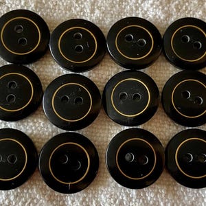 Könnte beinhalten: Zwölf schwarze Knöpfe mit jeweils zwei Löchern. Jeder Knopf hat einen goldfarbenen Ring am Rand. Die Knöpfe sind rund und scheinen aus glänzendem Material zu sein.