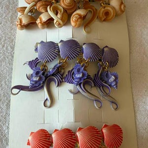 Pode incluir: Três ganchos de cabelo num cartão de apresentação branco com a etiqueta DISMAY. O gancho superior apresenta conchas em espiral castanhas, o do meio conchas roxas em leque e espiral, e o inferior conchas cor de laranja em leque e espiral.