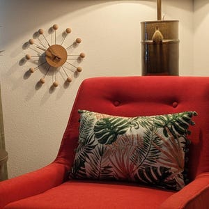 Könnte beinhalten: Innenraum im Mid-Century-Modern-Stil mit einem roten Sessel und einem Kissen mit tropischem Blättermuster. Eine Holzuhr mit Sonnenstrahlendesign hängt an der Wand. Eine Lampe und ein Beistelltisch sind ebenfalls zu sehen.