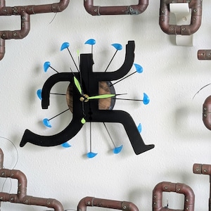 May include: A unique wall clock featuring a black running figure with blue accents and green clock hands. The clock is mounted on a white wall and surrounded by copper pipes, creating an industrial design.