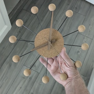 Puede incluir: Un reloj de pared moderno con esfera de corcho y manecillas de madera. El reloj presenta varillas metálicas negras con esferas de corcho. Un elemento decorativo para el hogar, con un diámetro de unos 30 cm.