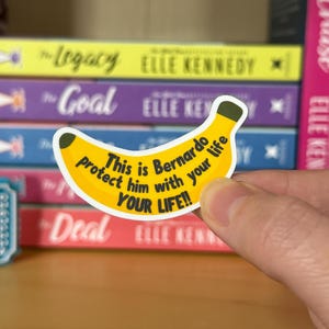 May include: A banana-shaped sticker with the text "This is Bernardo protect him with your life YOUR LIFE!!" in black. The sticker is yellow with a green stem. Books are in the background.