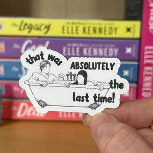 Puede incluir: Una pegatina blanca con una ilustración en blanco y negro de dos personas en una bañera. La pegatina tiene el texto "that was ABSOLUTELY the last time!" en una fuente manuscrita. La pegatina se sostiene frente a una pila de libros.